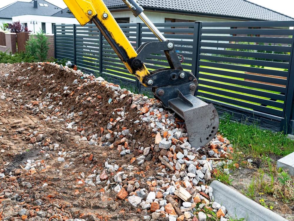 Koparka wykonująca prace ziemne przy usuwaniu gruzu i ziemi obok ogrodzenia posesji
