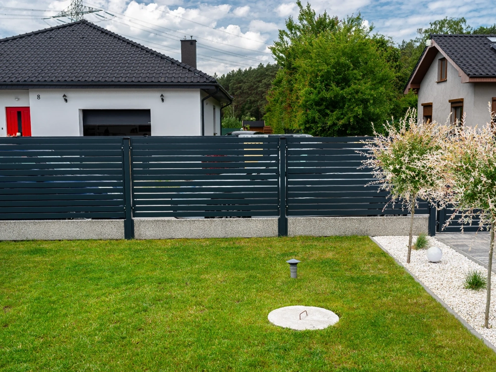 Ogrodzenia metalowe z poziomymi lamelami i betonową podmurówką przed domem, zadbany trawnik