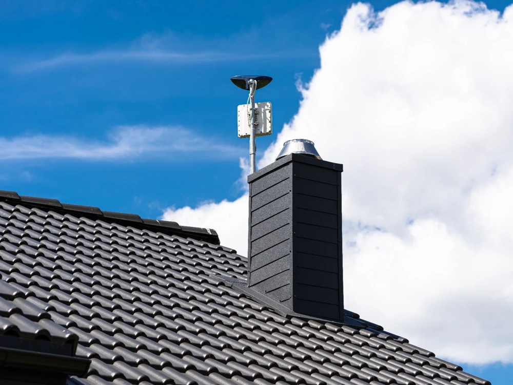 Obudowany komin z metalową czapą na pokryciu dachowym z czarnych dachówek