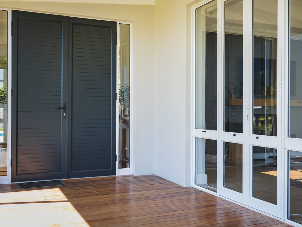 Wejście budynku z czarnymi drzwiami wejściowymi i białymi oknami tarasowymi na tle drewnianego tarasu – montaż drzwi