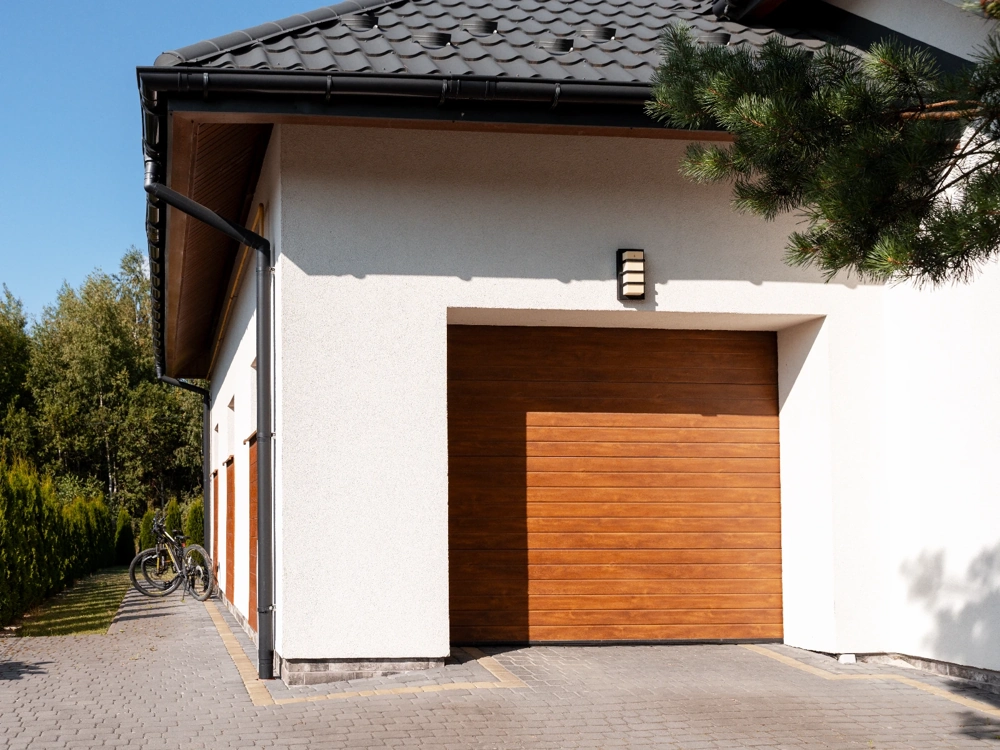 Drewniana brama garażowa zamontowana w białej elewacji z podjazdem z kostki brukowej