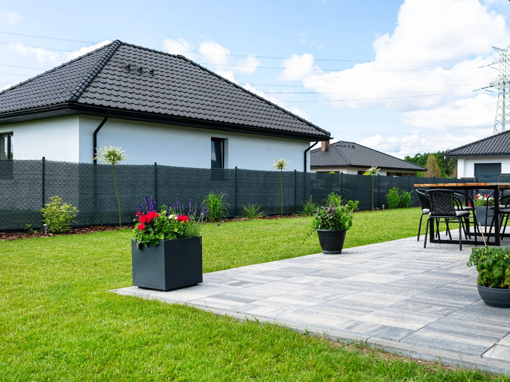 Ogrodzenia panelowe wokół tarasu z szarych płyt, donicami z kwiatami i trawnikiem przed nowoczesnym białym domem