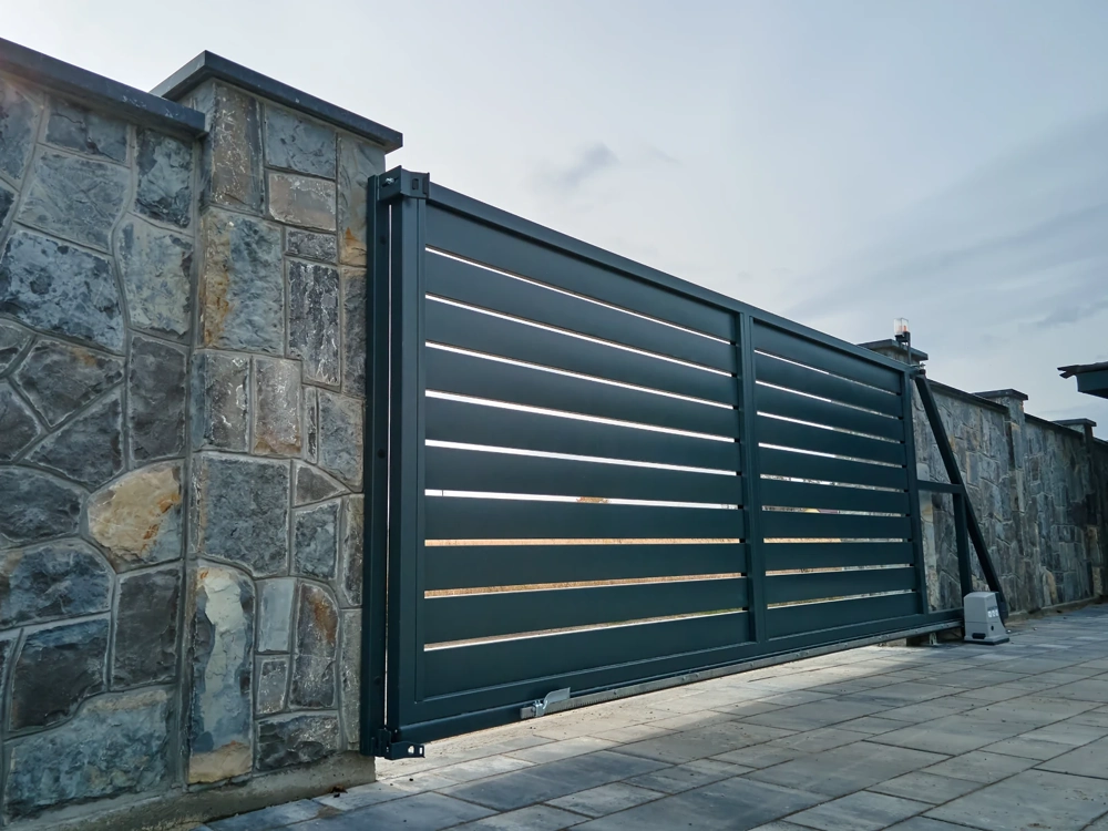 Przesuwna brama wjazdowa z metalowych paneli osadzona w kamiennym ogrodzeniu na podjeździe z kostki